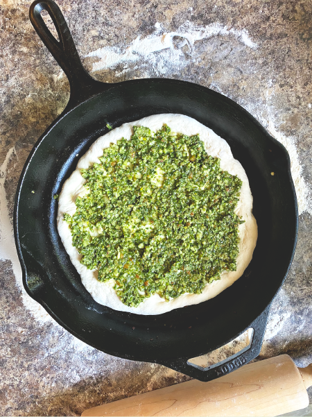 Pizza dough topped with green garlic pesto in a cast-iron skillet, almost ready to go in the oven!