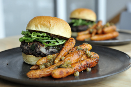 Black bean burger on a bun with arugula next to glazed carrots on a black ceramic plate.