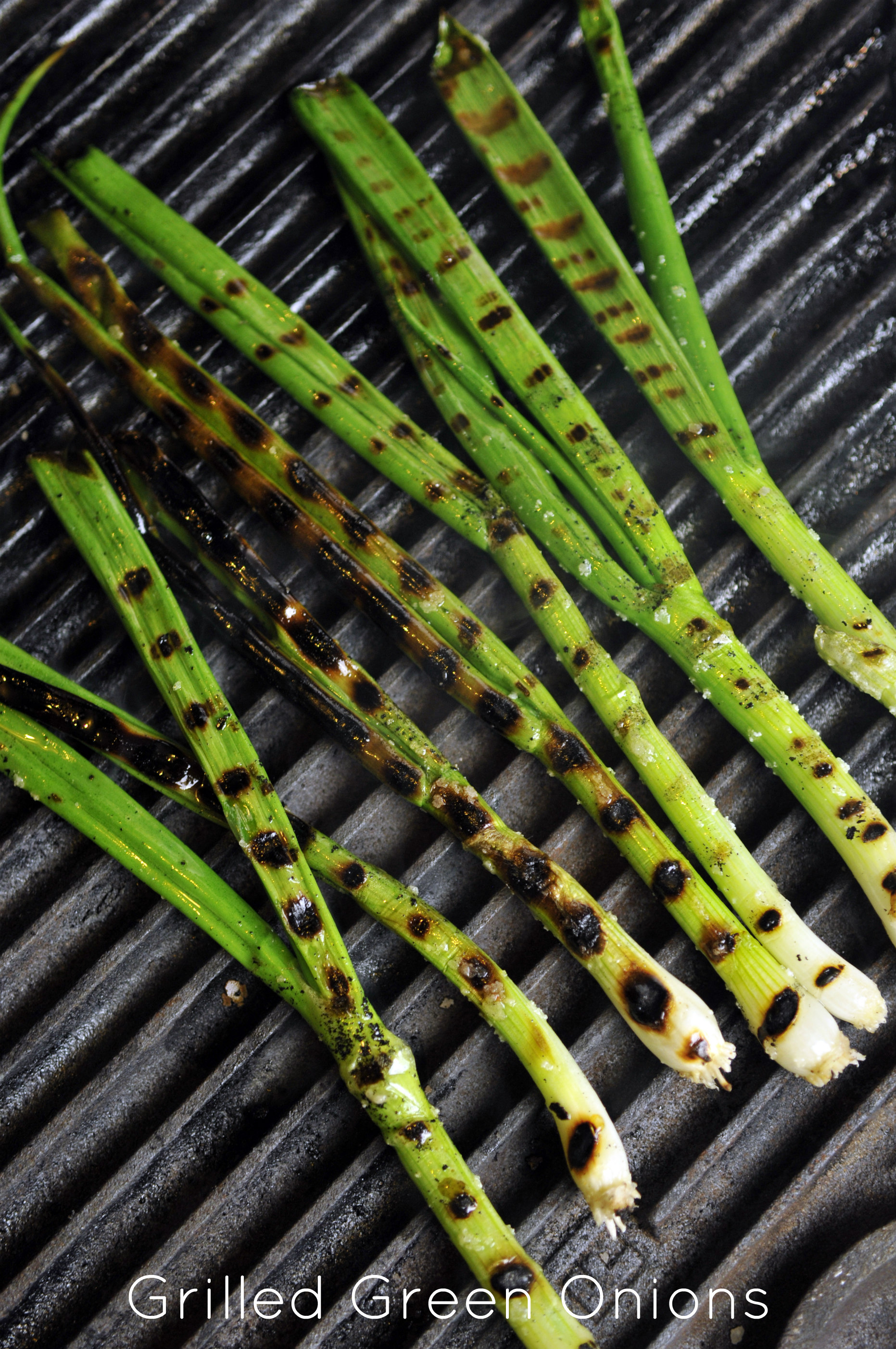 Seven skinny green scallions with white bulbs lay on a cast iron grill; they have amber colored grill marks.