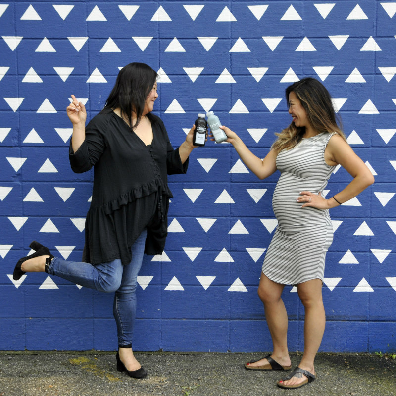 Joyce Cullen and Jo Um cheers with their juices in front of a bright blue wall with white triangles painted on it.
